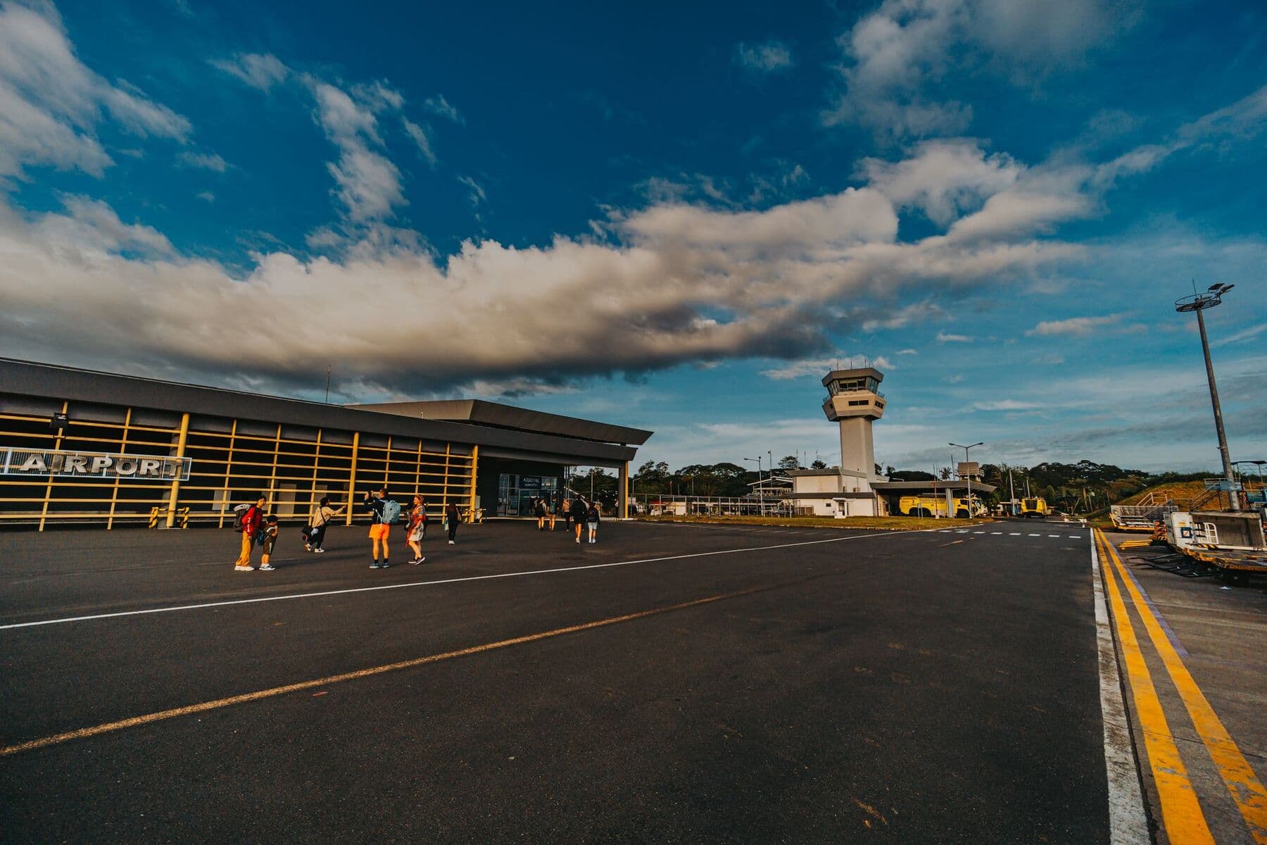 A small airport terminal and control tower beside a taxiway, with a few people walking on the apron under a blue sky.