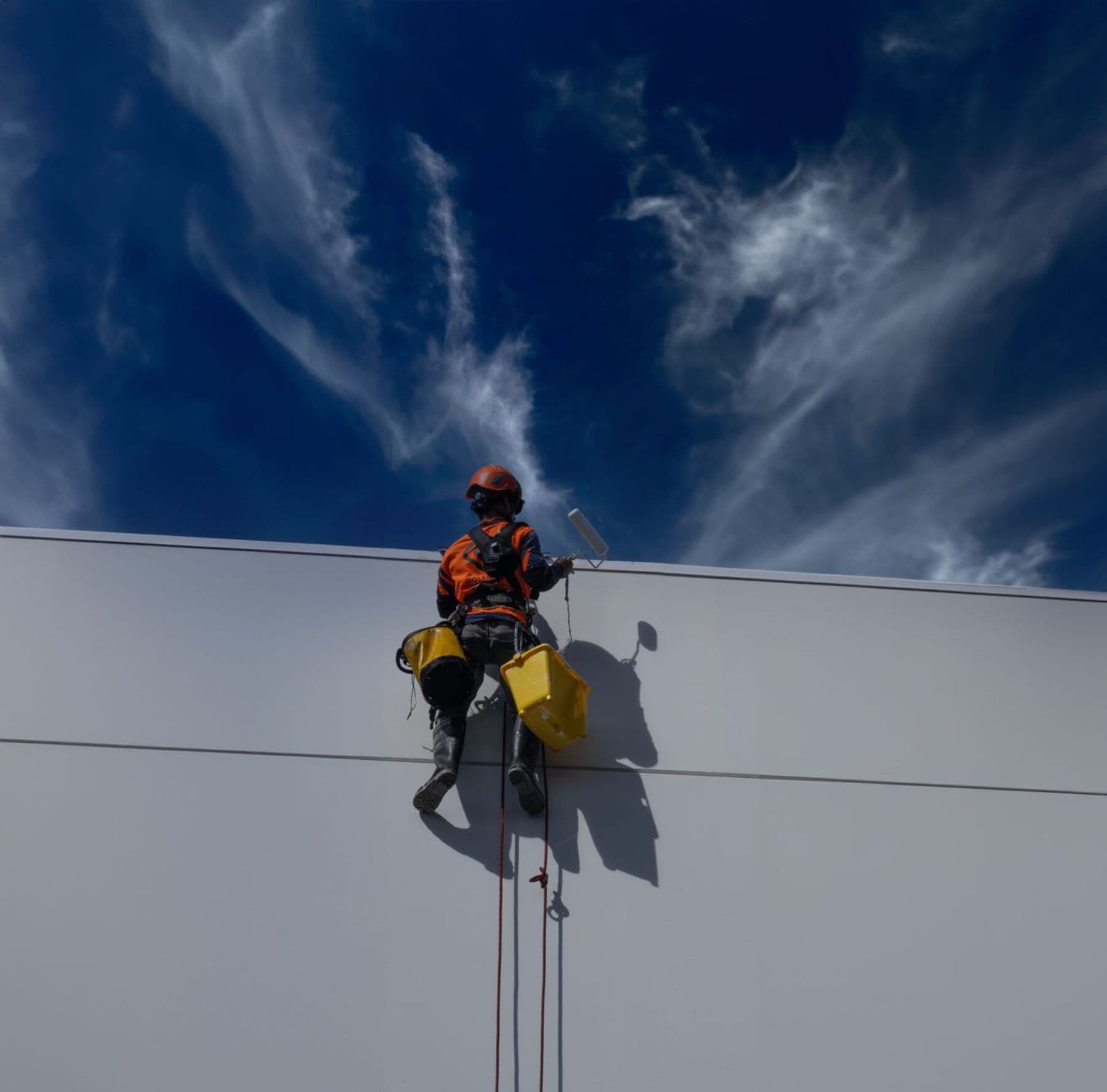 A worker in an orange helmet hanging from ropes on a large gray-white wall, with deep blue sky above.