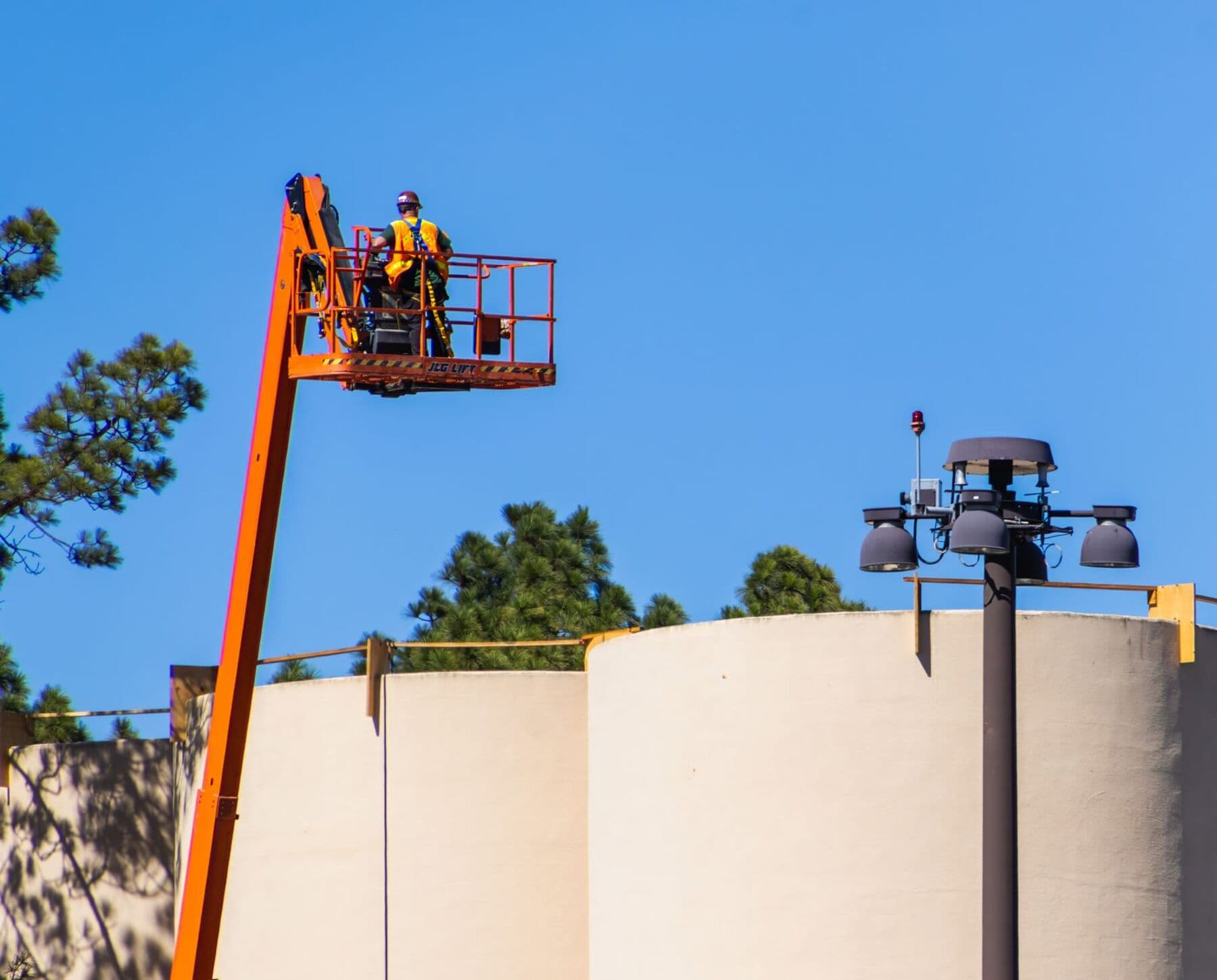 A worker in a high-visibility vest standing in the basket of an orange boom lift, raised beside a cream-colored wall under a clear blue sky.