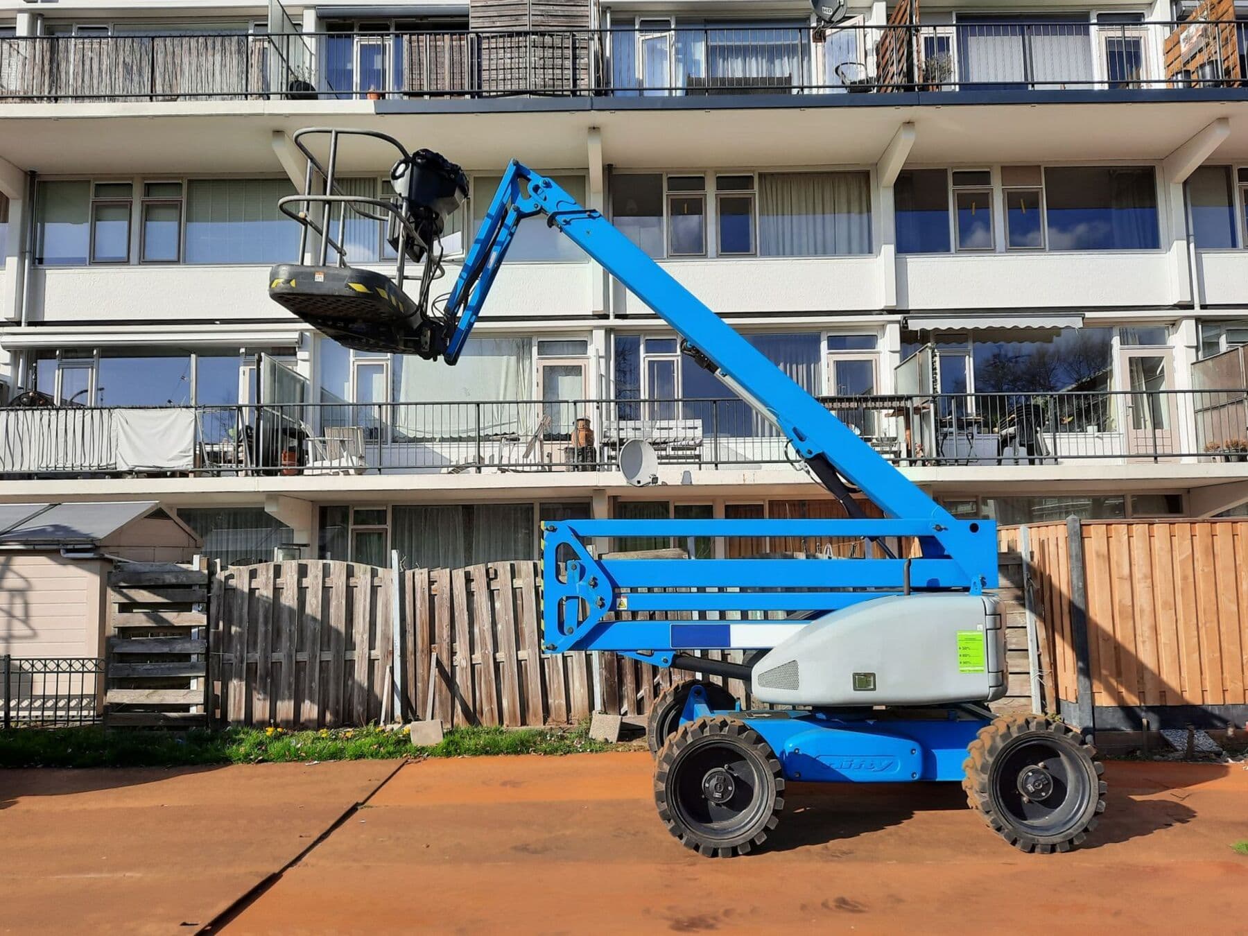 A blue boom lift parked on a dirt lot in front of a white residential building with long balconies.