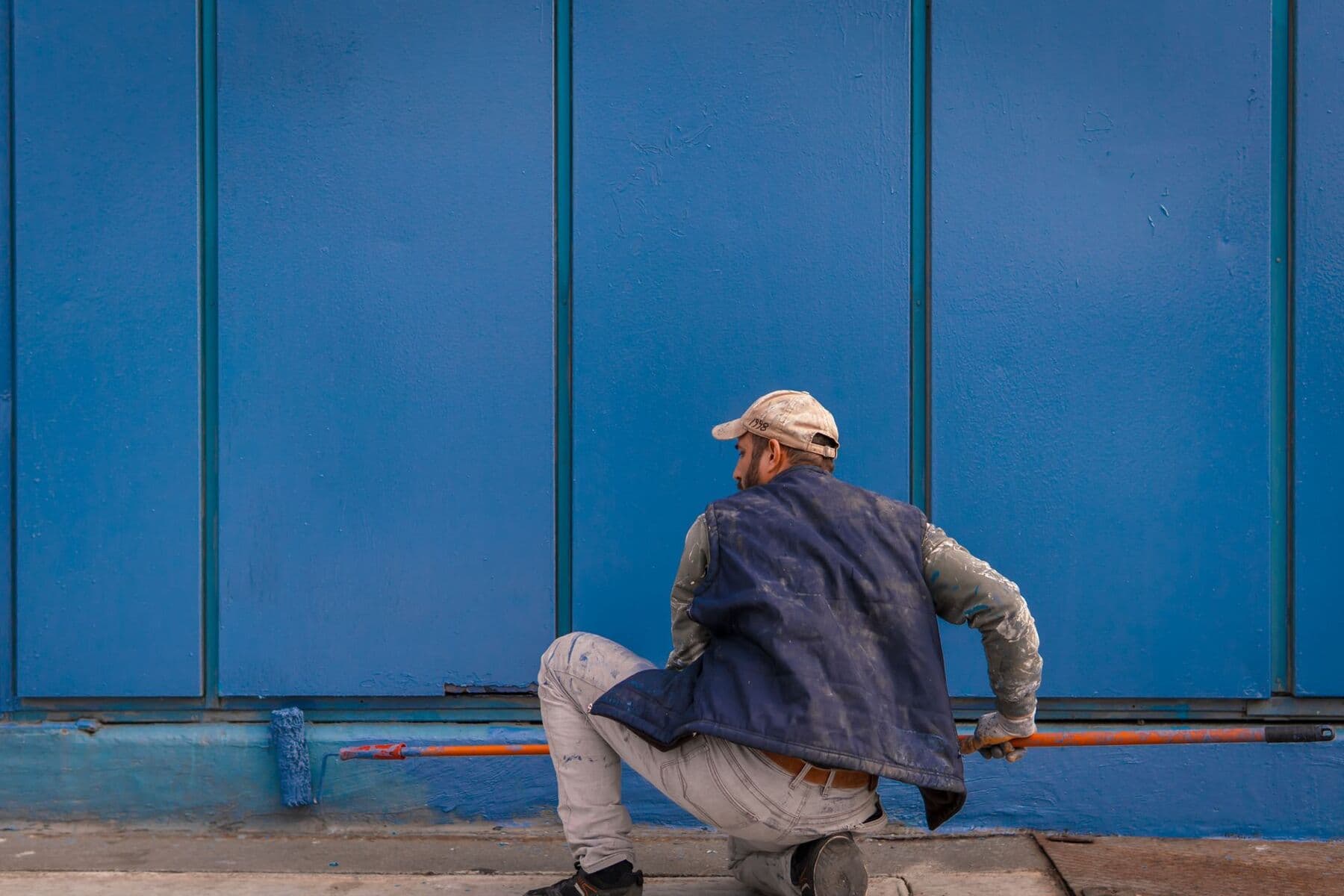 A worker crouching with a roller on a long pole, painting the base of a large blue wall.