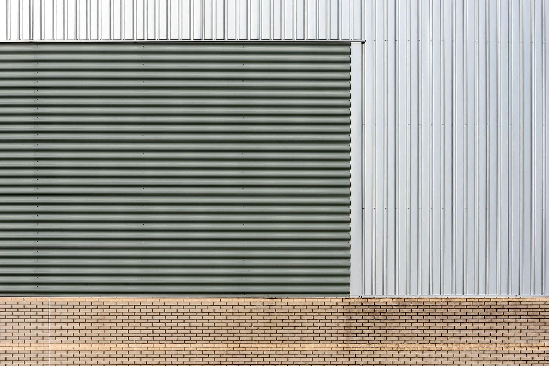 The wall of an industrial building clad in gray and silver metal panels over a brick base.